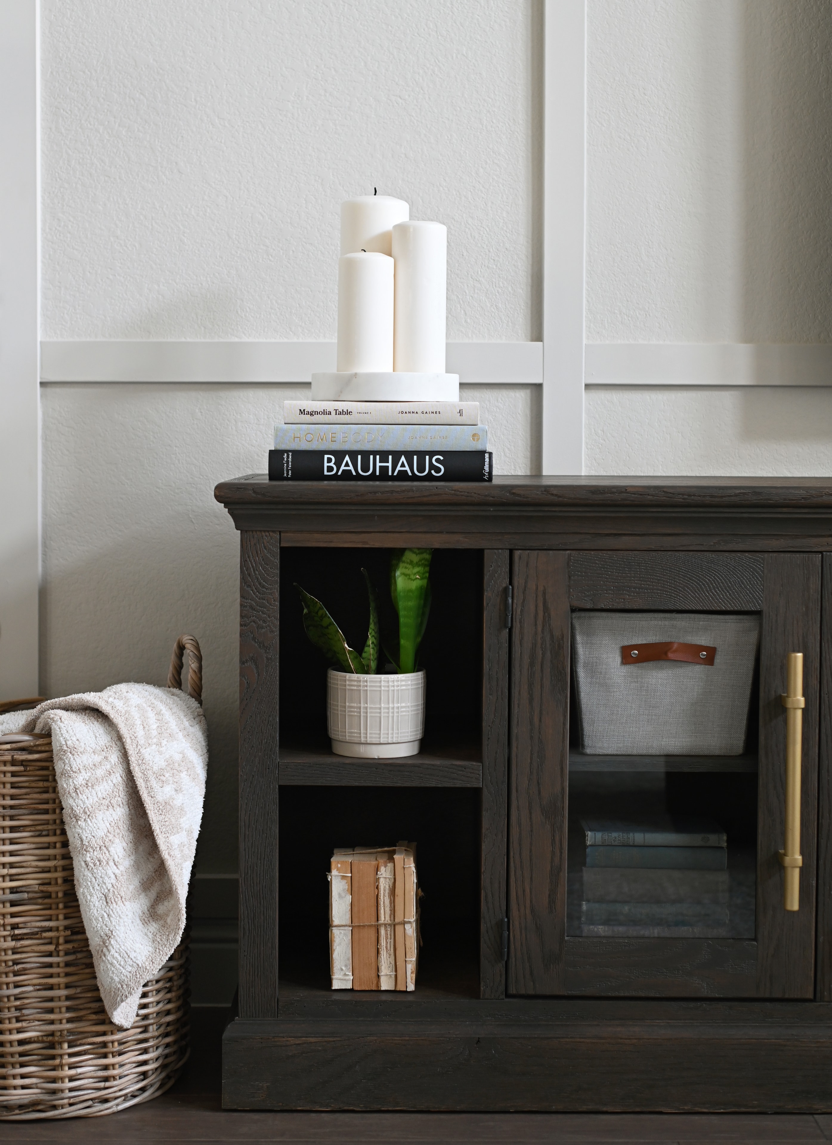 Dark wood cabinet styled with design books, candles, snake plant, and woven basket
