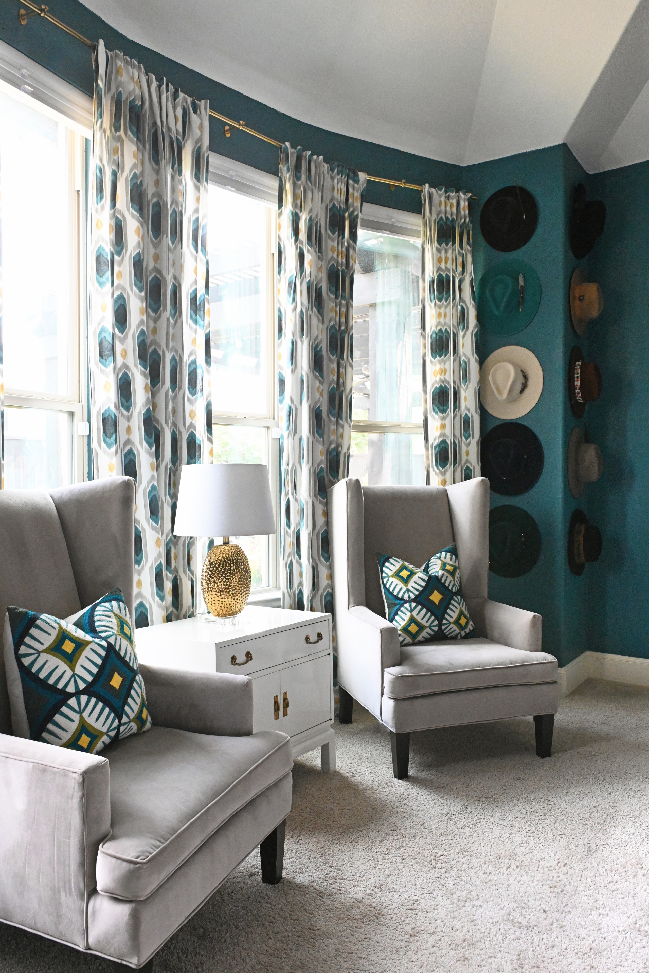 Sitting room with teal walls, patterned curtains, gray wingback chairs, and hat wall display